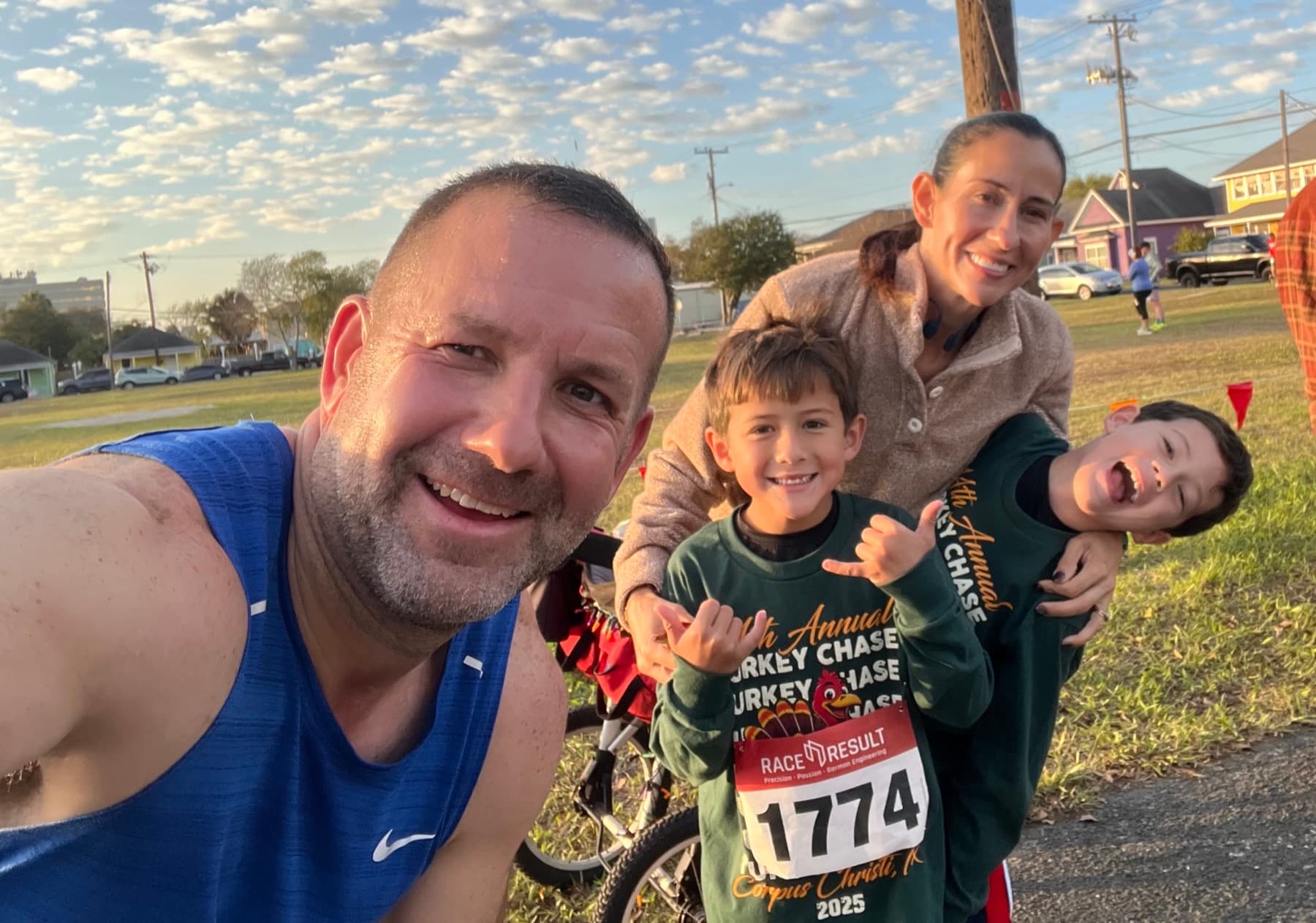 Will Bonneau taking a selfie with Alamar and their twin boys at the Turkey Chase race in Corpus Christi, one of the boys wearing race bib 1774.