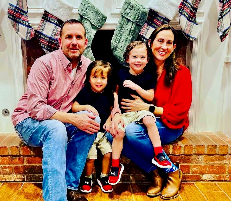 Alamar Cavada and Will Bonneau sitting on a brick hearth with their twin boys, in front of a fireplace hung with Christmas stockings.