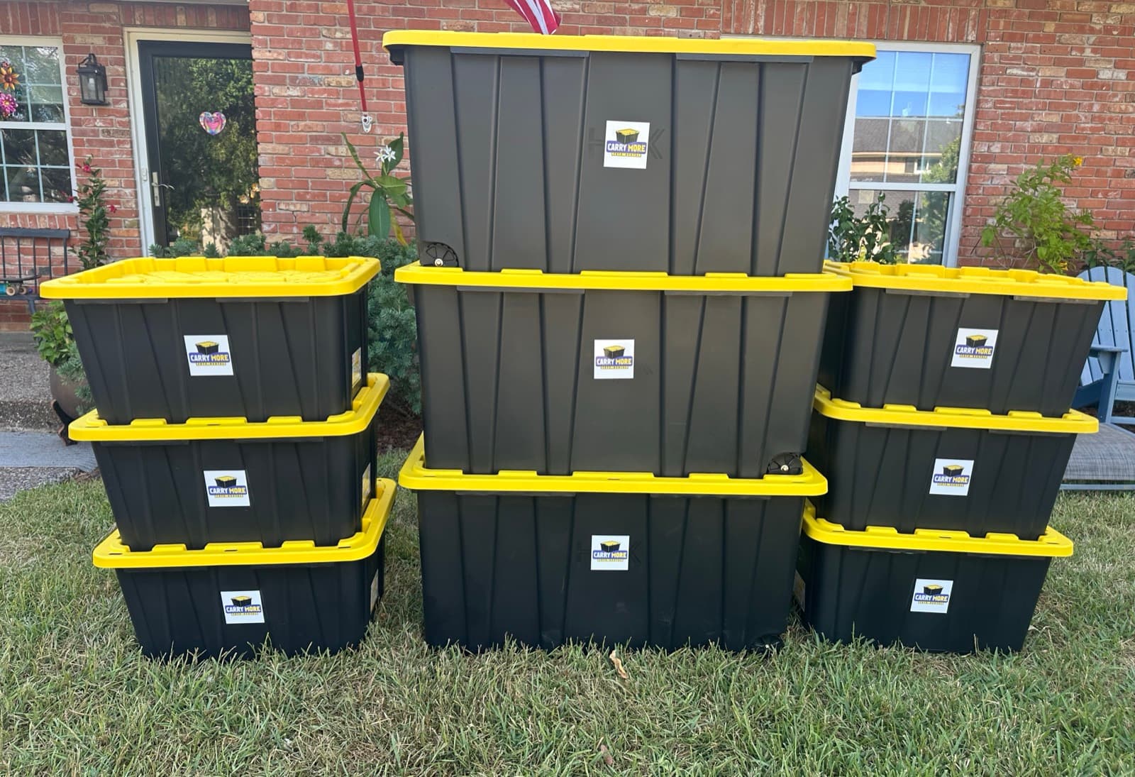 Nine black CarryMore totes with yellow lids, stacked three high in three columns on the grass in front of a brick house.